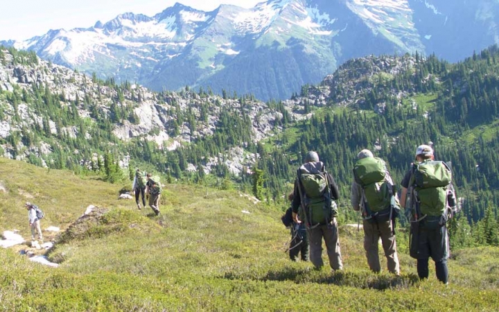 a group of outward bound students wearing backpacks hike along a grassy alpine area. There are vast mountains in the distance.